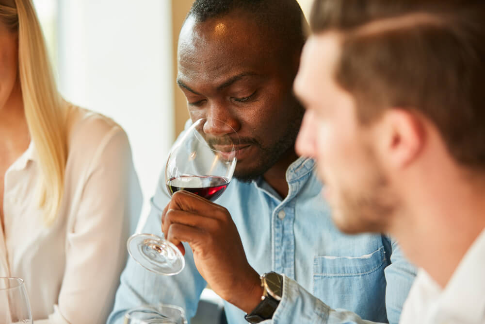 Na foto, um homem negro sente o aroma de um vinho. Ele está de camiseta azul e utiliza um relógio no punho.
