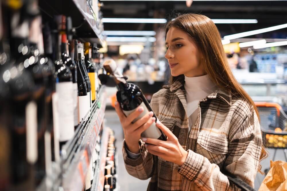 Imagem De Uma Mulher Lendo O Rótulo De Um Vinho Em Um Supermercado.