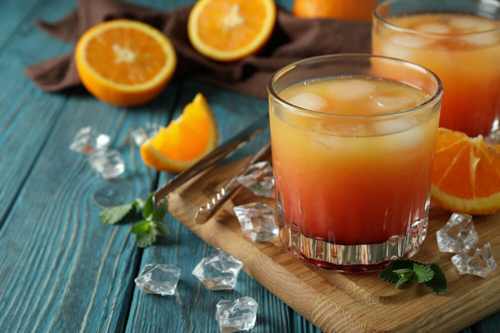 Imagem de uma Tequila Sunrise. Ao fundo, há laranjas cortadas ao meio.