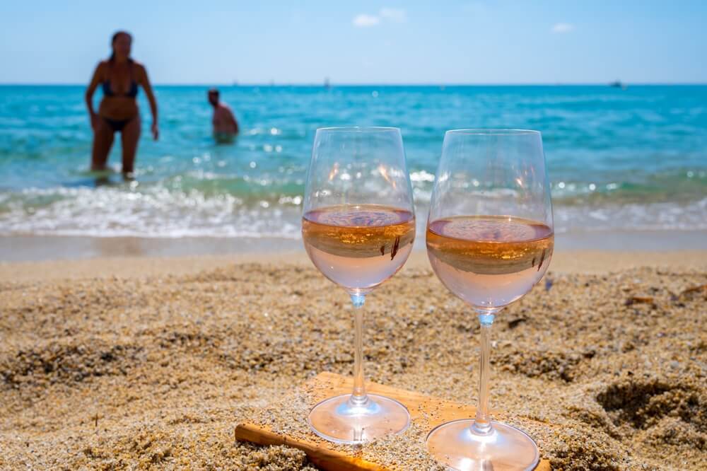 Imagem De Duas Taças De Vinho Rosé Na Praia. Ao Fundo, é Possível Notar O Mar Azul E Pessoas Brincando Na água.