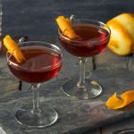 Duas taças com o drink boulevardier. Ao lado, há uma laranja descascada.