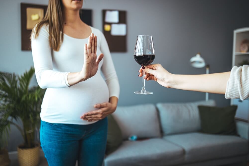 Imagem de uma mulher grávida, em pé, com uma mão na barriga e a outra recusando uma taça de vinho com álcool.
