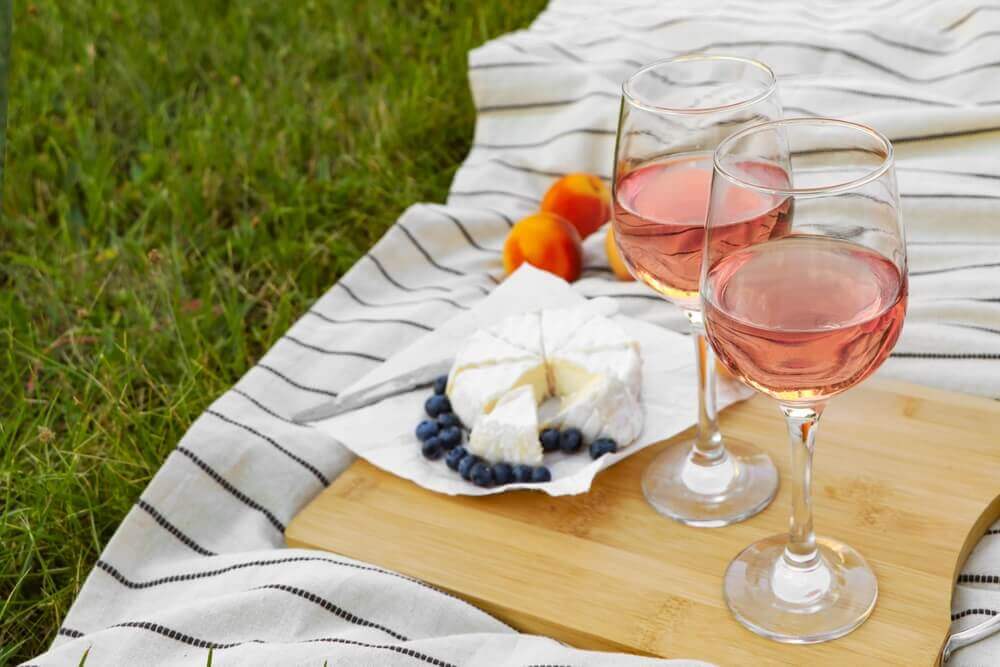 Imagem de duas taças de vinho rosé e, ao lado, um queijo repartido.