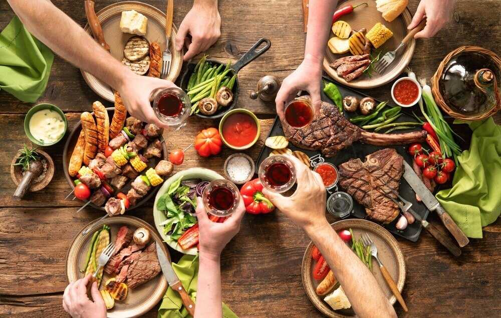 Imagem de uma mesa com diversos pratos com carne, além de legumes e vegetais. As pessoas estão fazendo um brinde com vinho..