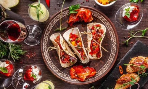 Imagem De Três Tacos Representando A Culinária Mexicano, Ao Lado De Outras Comidas. Há Ainda Uma Taça De Vinho.