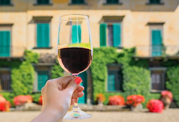 Pessoa segurando uma taça com vinho supertoscano e uma vinícola ao fundo, na região de Chianti