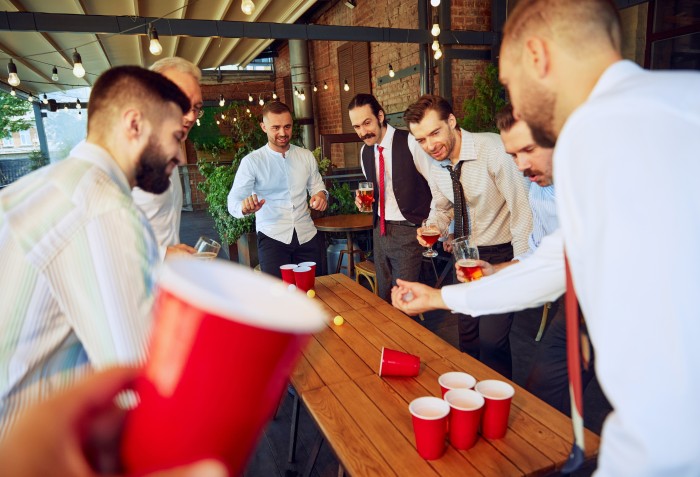 Alguns homens sorrindo e em pé tentando acertar bolinhas em copos que estão em cima da mesa