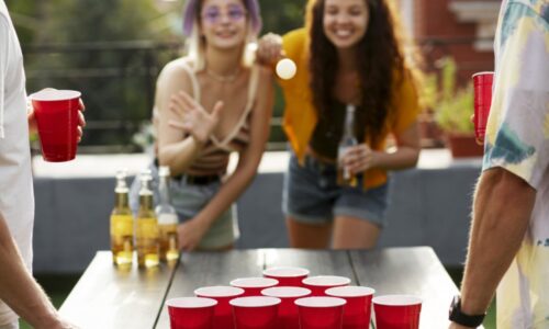 Dois Homens De Costas E Duas Mulheres De Frente Brincando Com Um Jogo De Bolinha Para Acertar Nos Copos