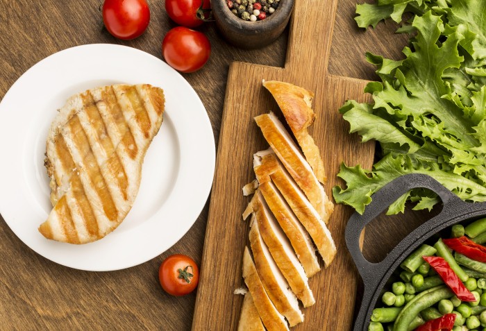 Pedaço de peito de frango grelhado em cima de um prato e pedaços cortados em cima de uma tábua com legumes ao redor