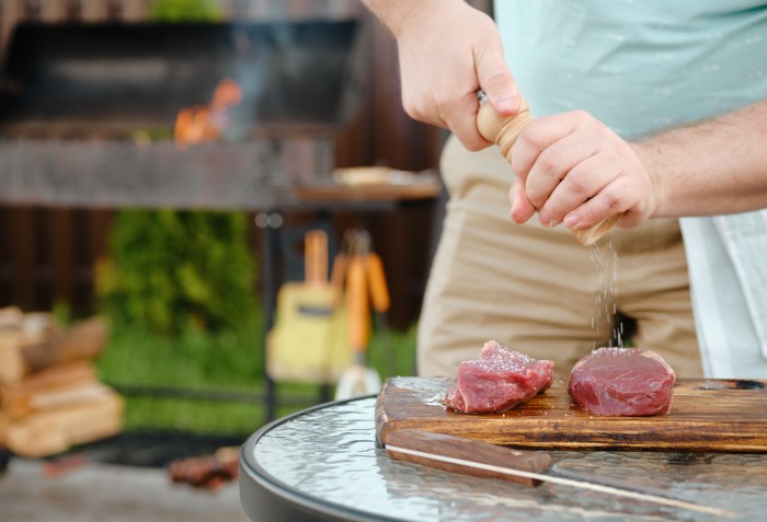 Mãos de um homem despejando sal em uma carne crua que está em cima de uma tábua de carne.