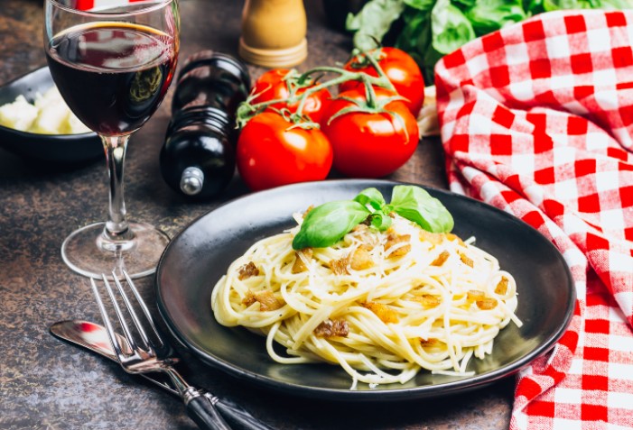 Macarrão a carbonara em um prato preto com taça de vinho e tomates ao redor.