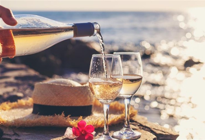 Alguém servindo duas taças de vinho branco, em uma paisagem de praia ao fundo.